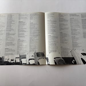 May include: Open pages of a vintage album featuring song lyrics and a black and white photograph of a row of classic cars. The cars are angled, creating a unique visual element. The background is a plain white surface.