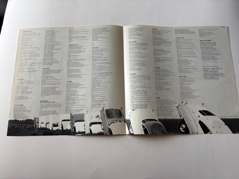 May include: Open pages of a vintage album featuring song lyrics and a black and white photograph of a row of classic cars. The cars are angled, creating a unique visual element. The background is a plain white surface.