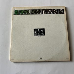 May include: A white vinyl record album with the title "The Hourglass" printed in black and green letters. The album cover features a black and white photo of a band.