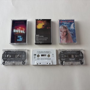 Pode incluir: Seis cassetes vintage com capas de álbuns. As cassetes incluem "Out of the Cellar" de Ratt, "Shot in the Dark" de Great White e outros álbuns. As caixas das cassetes são de plástico transparente e as capas dos álbuns apresentam várias cores e designs.