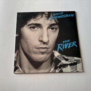 May include: Album cover for "The River" by Bruce Springsteen. The cover features a black and white portrait of Springsteen with the album title and artist name in blue text. The background is black.