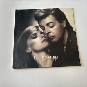 Pode incluir: Capa do álbum "Press to Play" de Paul McCartney. A capa apresenta uma imagem em preto e branco de um homem e uma mulher em um abraço próximo. O título do álbum e o nome do artista são impressos em branco.