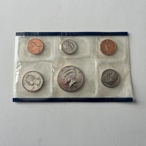 May include: A sealed coin set containing various U.S. coins, including a penny, nickel, dime, quarter, and half-dollar. The coins are encased in individual clear plastic pockets within a blue-bordered package. The half-dollar coin features a profile of John F. Kennedy.