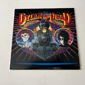 May include: A vintage vinyl record album cover featuring a colorful illustration of a train with the text "Dylan & The Dead" in a bold, stylized font. The cover features two figures, one with a red flower crown and the other with long dark hair.