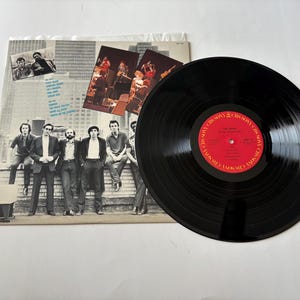 May include: A vinyl record album with a black disc and a red label. The album cover features a group of men in suits and casual wear. The album is on a white surface.
