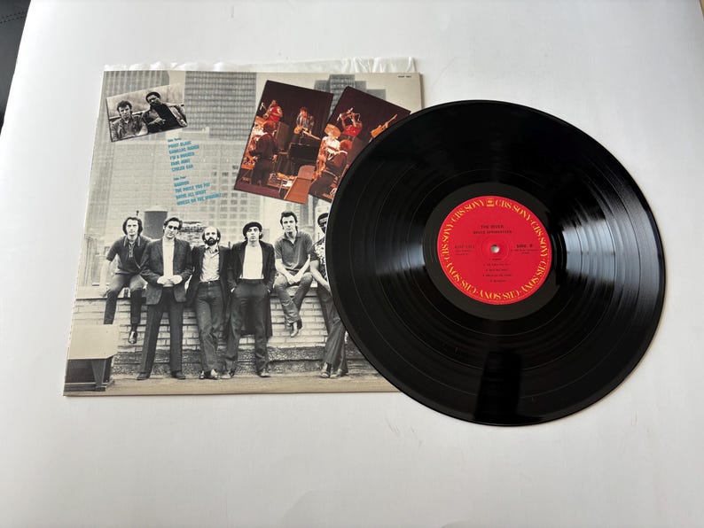May include: A vinyl record album with a black disc and a red label. The album cover features a group of men in suits and casual wear. The album is on a white surface.