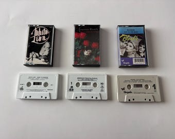 May include: Six vintage cassette tapes. Three are in their cases, featuring album art for "White Lion," "Concrete Blonde," and "Blondie." The other three tapes are visible, with labels. The tapes are white and gray.