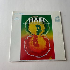 May include: A vintage vinyl record album cover for the musical "Hair: The American Tribal Love-Rock Musical". The cover features a psychedelic design with a person's face surrounded by a colorful, swirling pattern.