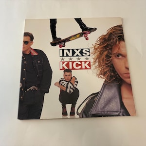 May include: Album cover for INXS's "Kick" featuring band members and a skateboard. The album title is in bold letters with stars. The cover has a white background and the band members are wearing black jackets.