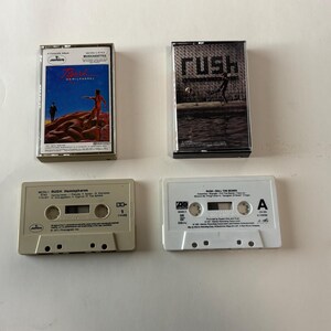May include: Four vintage RUSH cassette tapes. Two tapes are in their cases, featuring album art with the band's name. The other two tapes are visible, with labels and text. The tapes are in shades of gray and white.