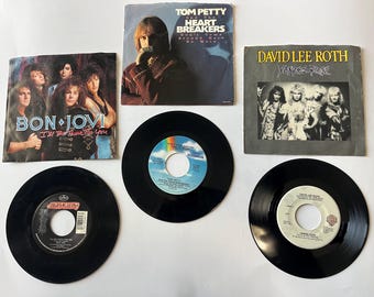 Puede incluir: Una colección de cinco discos de vinilo de 45 rpm. Los discos están en fundas negras con etiquetas blancas. Las etiquetas muestran los nombres de las bandas y los títulos de las canciones. Las bandas incluyen Bon Jovi, Tom Petty and the Heartbreakers, David Lee Roth y Van Halen.