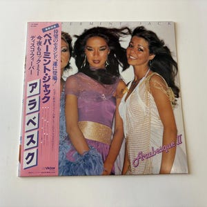 Peut inclure: Pochette d'album Arabesque II avec trois femmes en tenues glamour. La pochette de l'album contient du texte rose et blanc en japonais. Le titre de l'album est en écriture cursive.