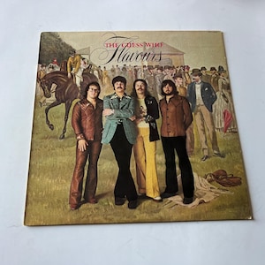 以下が含まれることがあります： 「ザ・ゲス・フー」というバンドの、ヴィンテージのビニールレコードアルバムのカバーで、アルバムのタイトルは「フレーバーズ」です。カバーアートは、1970年代の服装をした男性グループが、馬と騎手を背景に、野原に立っている様子を描いています。