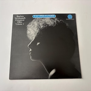 May include: A vintage vinyl record album cover for Barbra Streisand's Greatest Hits Volume 2. The cover features a black and white profile portrait of Streisand. The album title and artist name are printed in white. The words "Half Speed Mastered" are in a blue banner.