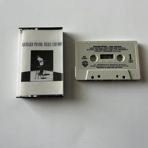 May include: A white cassette tape with the title "Richard Pryor - Here And Now" and a list of songs including "Here And Now", "Southern Hospitality", "Slavery", "Motherland", "Met The President", "Fire Exit" and "Musicoine (The One)".