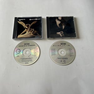 May include: Two compact discs, one with the title "Saints & Sinners" and the other with the title "Slide It In", both by Whitesnake. The discs are silver with black text and a small logo.