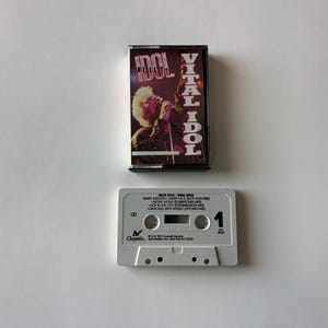 Puede incluir: Una cinta de cassette vintage y su estuche para el álbum "Vital Idol" de Billy Idol. El estuche de la cinta tiene un marco negro con una portada de álbum púrpura y dorada. La cinta de cassette es blanca con la lista de canciones.