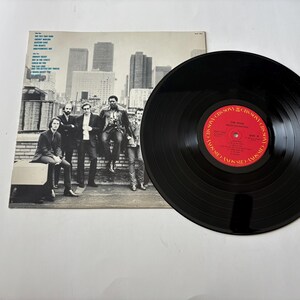 May include: A vintage vinyl record with a black surface and a red label. The album cover features a black and white photo of a group of men on a rooftop with a city skyline in the background. The record is a music collectible.