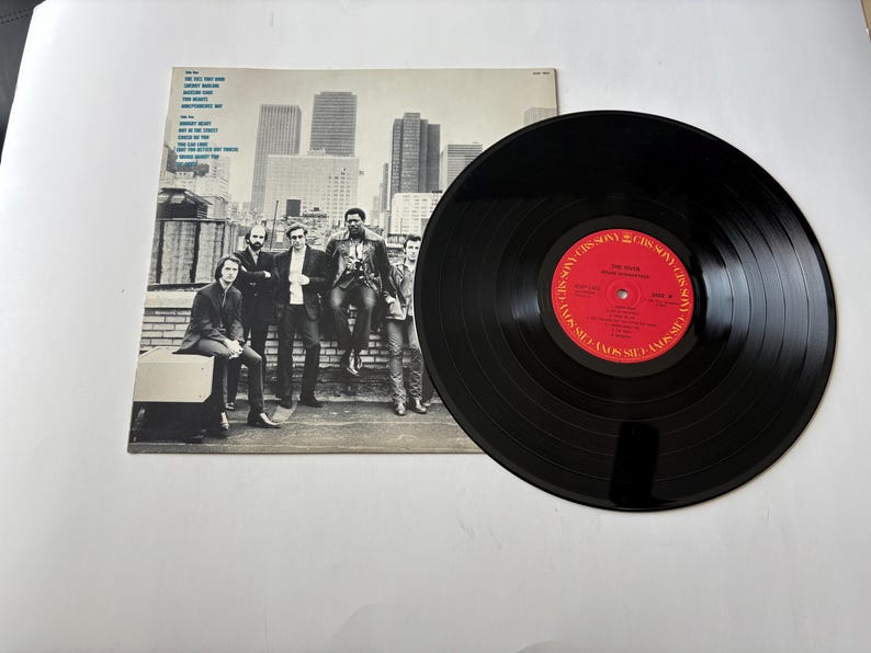 May include: A vintage vinyl record with a black surface and a red label. The album cover features a black and white photo of a group of men on a rooftop with a city skyline in the background. The record is a music collectible.