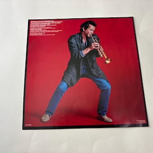 Puede incluir: Portada de &aacute;lbum con un hombre tocando la trompeta sobre un fondo rojo. Viste una chaqueta oscura, vaqueros azules y zapatos estampados. Texto visible en la parte superior izquierda.