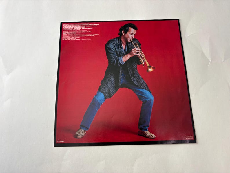 Puede incluir: Portada de &aacute;lbum con un hombre tocando la trompeta sobre un fondo rojo. Viste una chaqueta oscura, vaqueros azules y zapatos estampados. Texto visible en la parte superior izquierda.