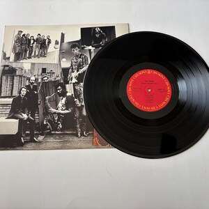 May include: A black vinyl record with a red label featuring text and a group of musicians on the album cover. The album cover is a collage of black and white photos of the band.