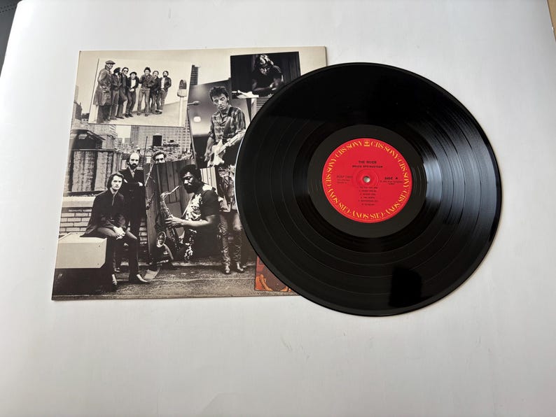May include: A black vinyl record with a red label featuring text and a group of musicians on the album cover. The album cover is a collage of black and white photos of the band.