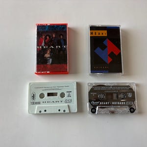 Op de afbeelding: Vier cassettebandjes worden getoond op een wit oppervlak. Twee bandjes tonen de band Heart, met albumhoezen met de bandleden. De andere twee bandjes hebben albumhoezen met geometrische ontwerpen.