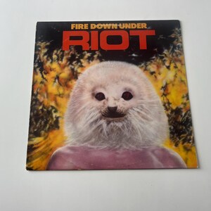 Może przedstawiać: Okładka płyty winylowej z białym stworzeniem przypominającym fokę, z różowym ciałem i żółto-brązowym tłem. Tytuł albumu to "FIRE DOWN UNDER RIOT" czerwonymi literami.
