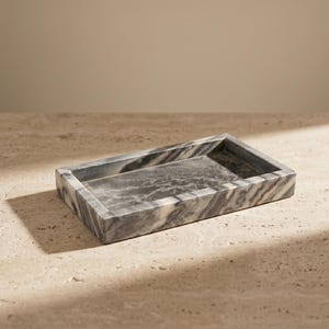 Peut inclure: Un plateau rectangulaire en marbre avec des veines grises et blanches. Le plateau a un profil bas et un bord légèrement surélevé. La surface est lisse et polie, adaptée pour contenir de petits objets. Le plateau est sur une surface claire.