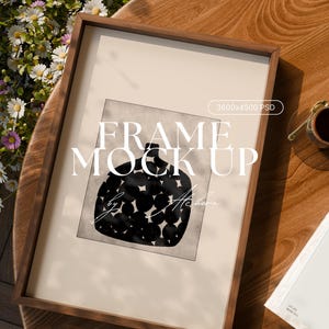 Puede incluir: Un marco de madera oscura que contiene una impresión artística. La obra de arte presenta un diseño abstracto en blanco y negro con forma de jarrón y patrones circulares. El texto "FRAME MOCK UP" se muestra en la obra. El marco está sobre una mesa de madera con flores.