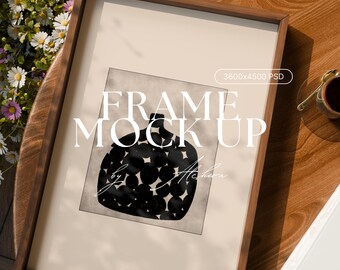 May include: A framed art print with a dark wood frame. The artwork features a black and white abstract design of a vase-like shape with circular patterns. The text "FRAME MOCK UP" is displayed on the artwork. The frame is on a wooden table with flowers.