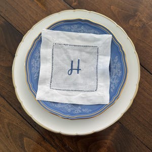 May include: A white linen napkin with a blue embroidered letter "H" sits atop a blue and white patterned plate, which is placed on a larger white plate with a gold rim. The plates are arranged on a dark wooden surface.