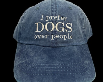 May include: A blue baseball cap with the embroidered phrase "I prefer DOGS over people" in white. The cap has a curved brim and an adjustable strap at the back. The hat is made of a soft, washed cotton material.