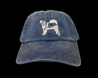 May include: A denim blue baseball cap featuring an embroidered image of a dog. The dog is depicted in white, black, and gray thread. The cap has a curved brim and an adjustable strap at the back.
