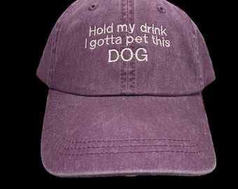 May include: A purple baseball cap with the embroidered text "Hold my drink I gotta pet this DOG". The cap has a curved brim and a classic six-panel design. The hat is made of a soft, washed fabric.