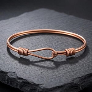 Puede incluir: Una pulsera de brazalete de color cobre con cierre de gancho. La pulsera está hecha de dos alambres paralelos que están envueltos en cada extremo y conectados con un lazo. La pulsera se muestra sobre una superficie oscura y texturizada.