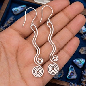 May include: A pair of silver earrings with a distinctive spiral design. The earrings have a wavy line leading to a spiral at the base. The earrings are shown on a hand.