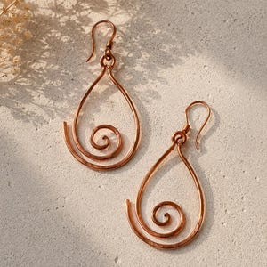 Puede incluir: Un par de pendientes de lágrima de color cobre con un diseño en espiral. Los pendientes tienen un cierre de gancho y se muestran sobre una superficie clara y texturizada. Los pendientes son una joya hecha a mano.