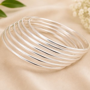 May include: A stack of silver bangles, each with a simple, rounded design. The bracelets are arranged on a beige, silky fabric, with green leaves and white flowers in the background. The bangles are reflective and shiny.