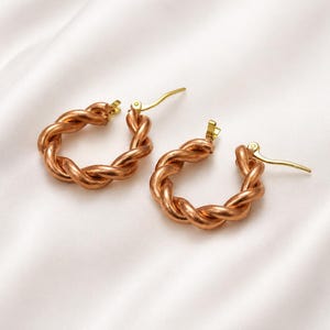 Könnte beinhalten: Ein Paar kupferfarbene Creolen mit einem gedrehten Seildesign. Die Ohrringe haben goldfarbene Verschlüsse und werden auf einer weißen, strukturierten Oberfläche präsentiert. Ein Schmuckstück.