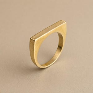 Könnte beinhalten: Ein goldfarbener Ring mit einer flachen, horizontalen Leiste an der Oberseite, die sich über die Breite des Bandes erstreckt. Die Metalloberfläche hat ein glattes, poliertes Finish mit subtilen Farbvariationen und bildet eine solide Struktur.