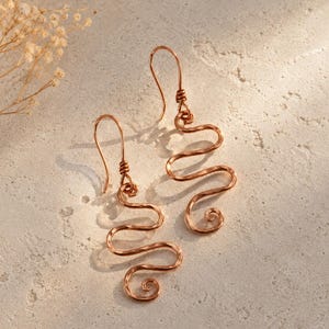 Può includere: Un paio di orecchini in filo di rame. Ogni orecchino presenta un lungo design ondulato con una spirale nella parte inferiore. Gli orecchini sono sospesi a un semplice gancio. Lo sfondo è una superficie neutra e strutturata.