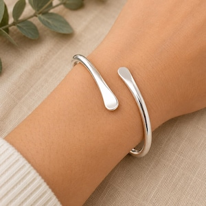 Peut inclure: Un bracelet manchette argenté avec une finition polie. Le design ouvert présente des extrémités incurvées et effilées. Le bracelet est porté sur un poignet. Le métal est brillant et réfléchissant.