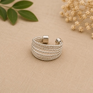 Op de afbeelding: Een zilverkleurige manchetring met een multi-strengs touwontwerp. De ring heeft een opening aan de bovenkant en wordt gepresenteerd op een beige oppervlak. De ring is een sieraad.
