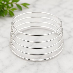 Può includere: Una pila di cinque braccialetti d'argento. I braccialetti sono sottili e rotondi, con una superficie lucida e riflettente. I gioielli sono esposti su una superficie di marmo bianco, con una pianta verde sfocata sullo sfondo.