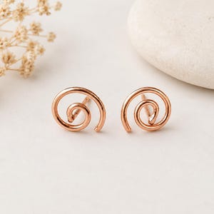 May include: A pair of rose gold spiral stud earrings. The earrings are made of thin, curved metal, forming a spiral shape. The earrings are displayed on a white surface, with a white stone and dried flowers in the background.