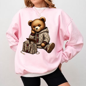 May include: A light pink sweatshirt featuring a brown teddy bear wearing a patterned hoodie and matching shoes. The bear is seated next to a matching handbag. The design has a luxury aesthetic.