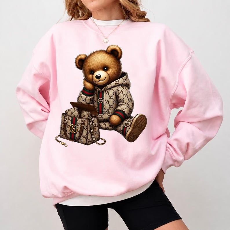 May include: A light pink sweatshirt featuring a brown teddy bear wearing a patterned hoodie and matching shoes. The bear is seated next to a matching handbag. The design has a luxury aesthetic.