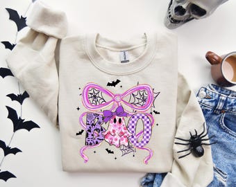 Może przedstawiać: Beżowa bluza z motywem Halloween. Projekt przedstawia słowo "BOO" z duchem i kokardą, ozdobione pajęczynami i nietoperzami. Projekt zawiera kolory różowy, fioletowy i czarny.
